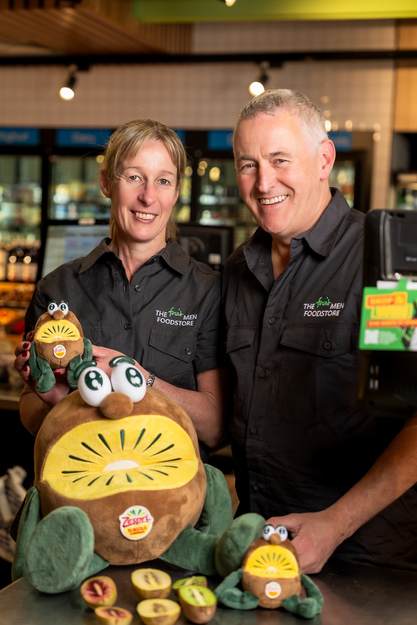 Two people in black uniforms holding a plush kiwi toy in a store setting.