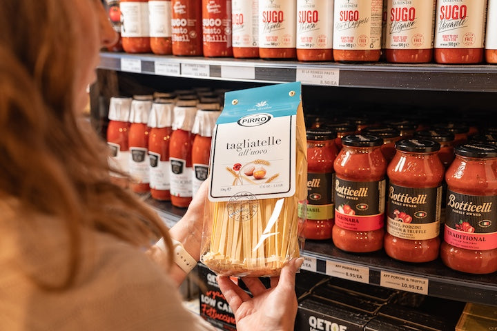 Person holding a package of pasta in front of shelves with pasta sauce bottles.
