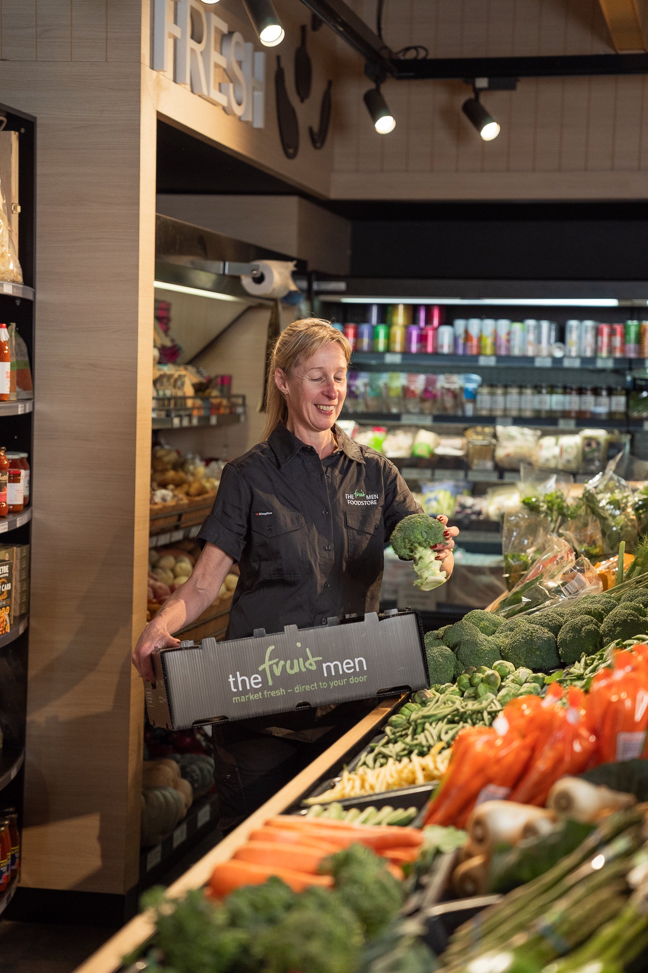 Person in a grocery store holding a box labeled 'the fruit men' with vegetables in the background.